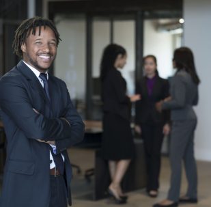 Portrait of businessman, arms folded, smiling, businesswomen talking in background