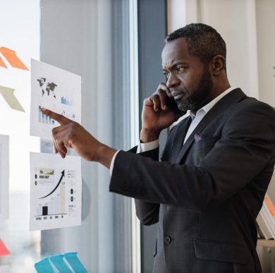 Focused multiracial businessman in smart suit receiving phone call while standing near glass panel with graphs and charts in corner office. Senior executive listening to worker's speech over mobile.
